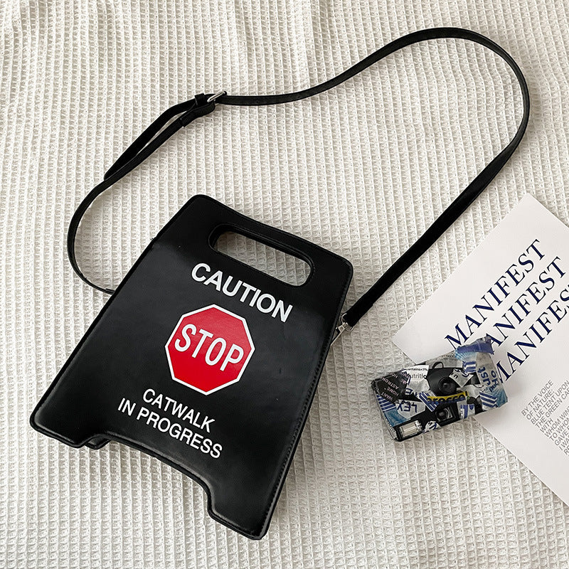 Black bag with 'Caution: Stop, Catwalk in Progress' text and a red stop sign on a textured white surface.