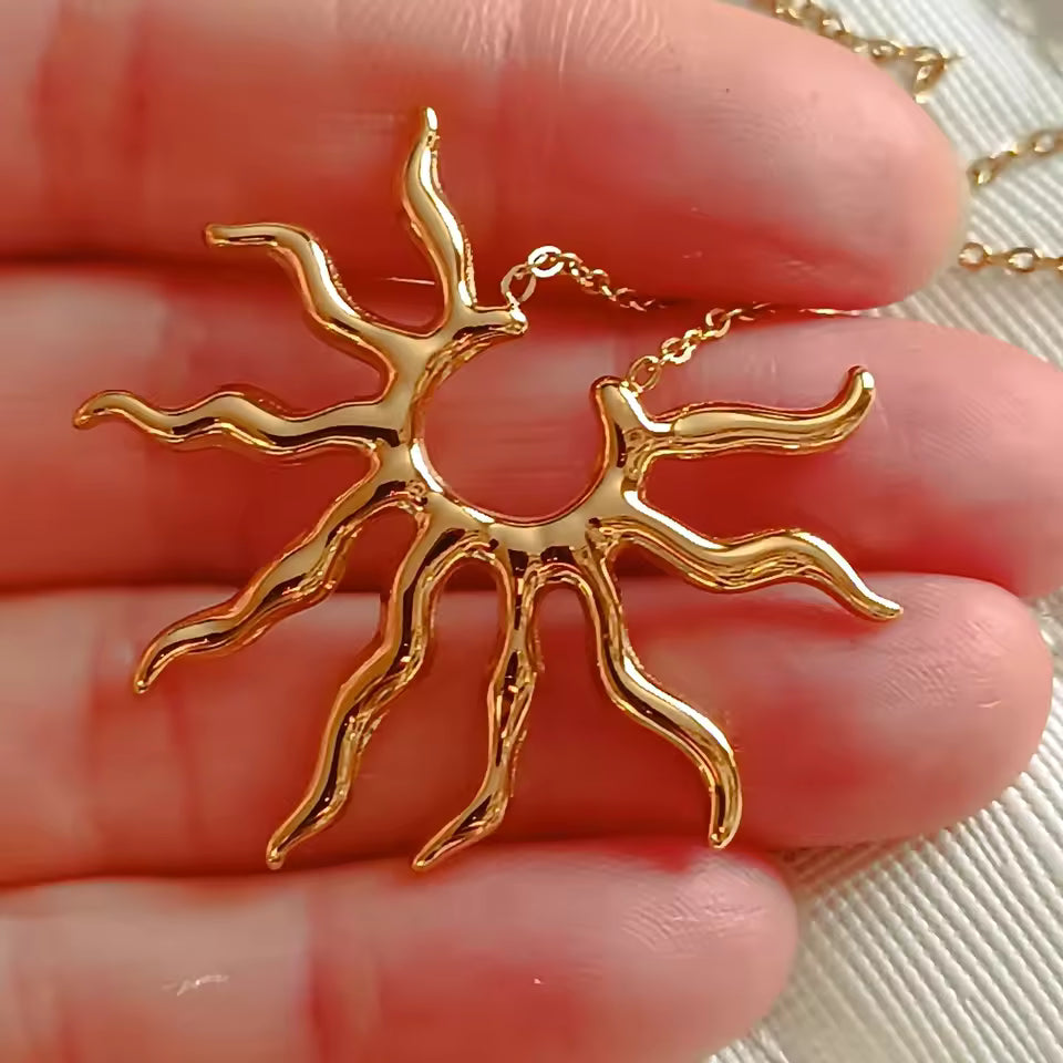 A gold-toned sun pendant necklace with a textured, organic branch-like design, held in a person's fingers against a light fabric background.